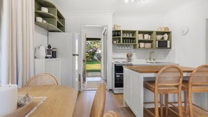 A kitchen with white cabinets, a wooden dining table, wicker chairs, and a view of the backyard. - Piccolo Cottage - Firepit - 4 Mins Walk to Beach (Callala Beach)