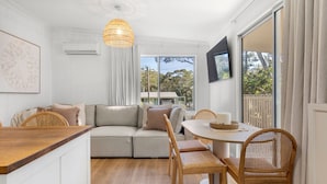 A modern living room with a sofa, dining table, and chairs. There is a large window with a view of trees and a house. - Piccolo Cottage - Firepit - 4 Mins Walk to Beach (Callala Beach)