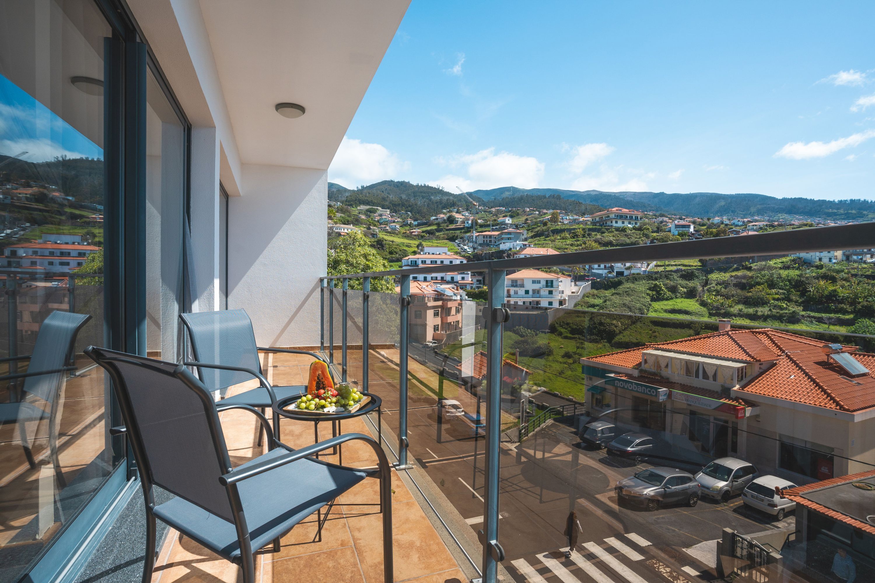 A balcony with a table and chairs, overlooking a town with buildings and greenery.