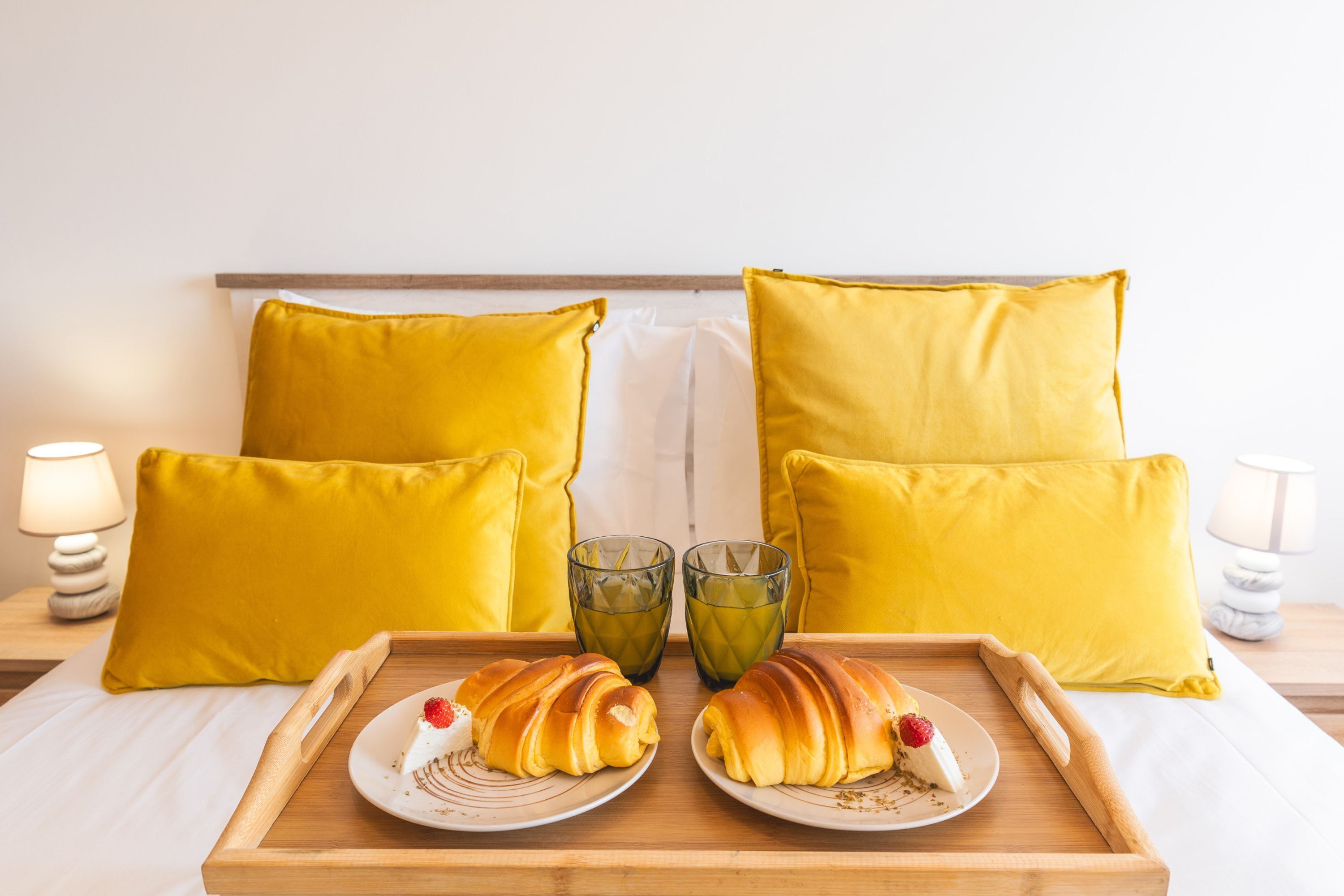 A breakfast tray with croissants and a strawberry on a bed with yellow pillows and two glasses of green juice.