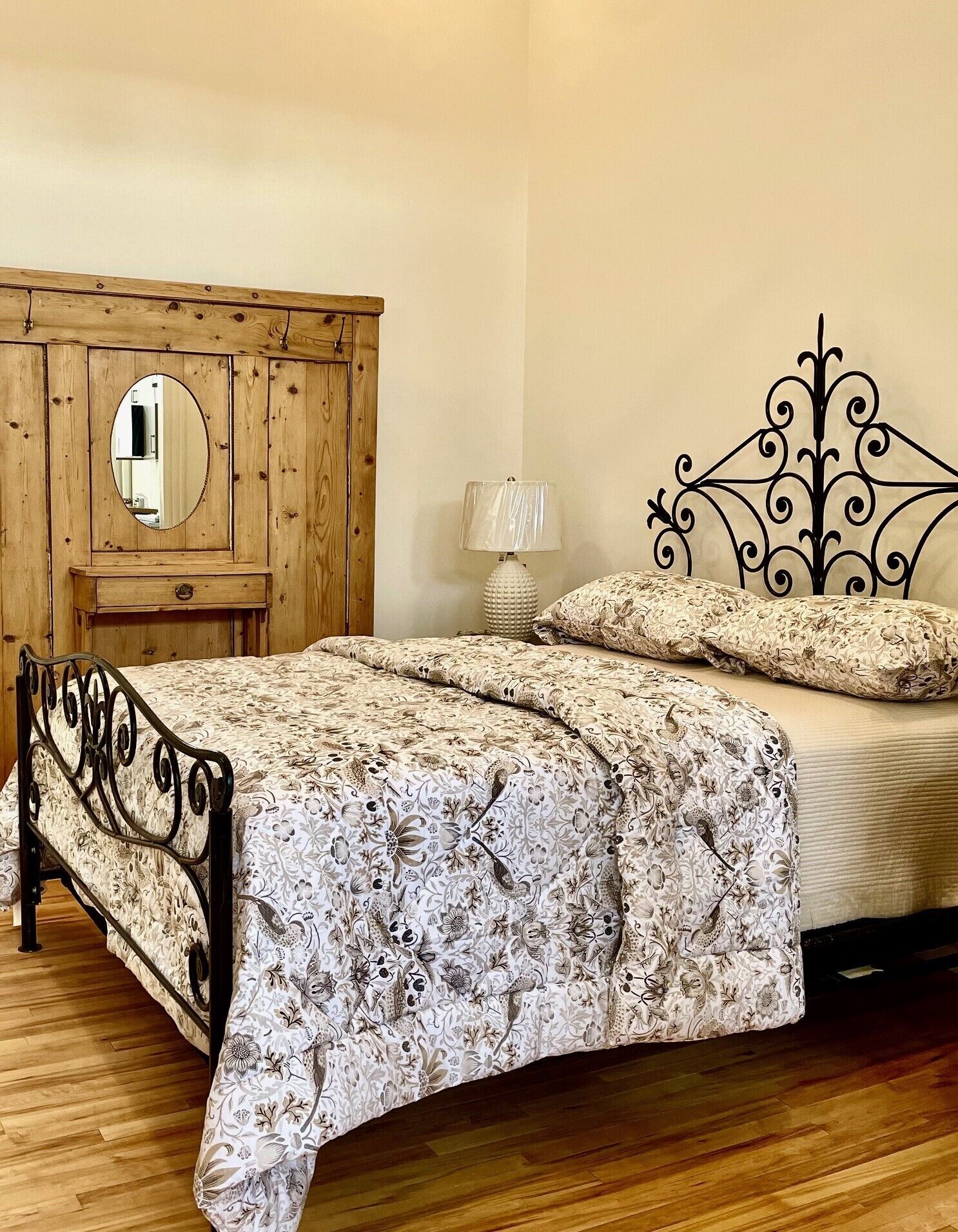 A bedroom with a wooden wardrobe, a bed with a floral bedspread, a bedside lamp, and a decorative mirror.
