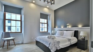 Une chambre à coucher moderne avec un grand lit, deux fenêtres, une chaise et une table de chevet avec une lampe.