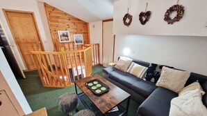 Un salon chaleureux avec un canapé foncé, une table basse en bois et deux poufs moelleux. Un escalier en bois mène à un niveau supérieur.