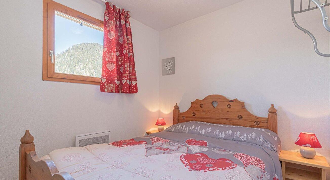 Quarto com cama de madeira, duas luminárias de cabeceira, janela com vista para uma floresta e cortina vermelha com padrão de coração.