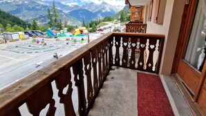 A balcony with a view of mountains, a parking area with colorful play equipment, and a red carpet leading to a door. - 50m from ski slopes, 2nd floor, view mountain, balcony, tv, ski locker, 23m², Valfréjus (Valfréjus)