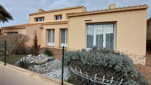 A two-story building with a beige exterior and a tiled roof. There are multiple windows, some with curtains, and a large sliding glass door. The building is surrounded by a wire fence and has a small garden area with rocks and shrubs.