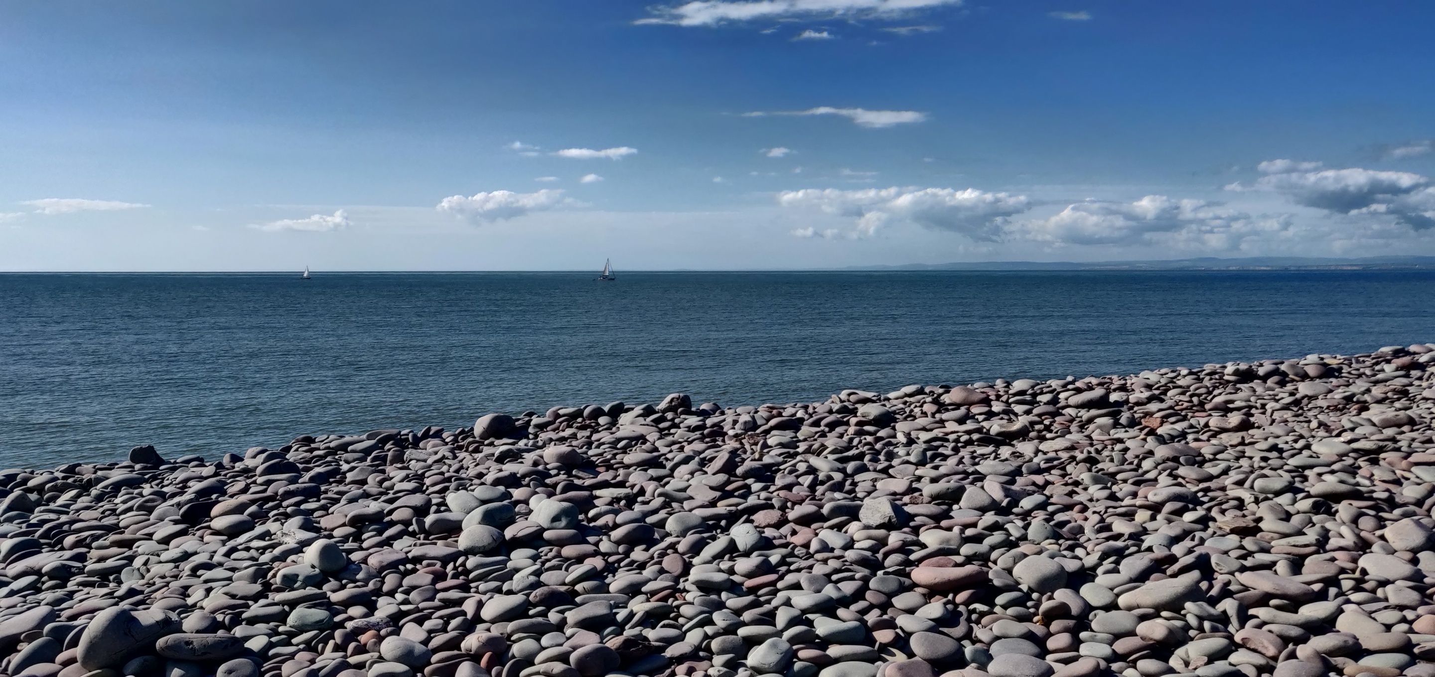 A pebbled beach with a sailboat in the distance.