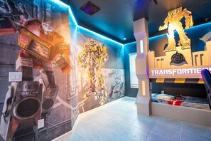 Una habitación con un amplio mural con temática de Transformers, un escritorio con computadora y una ventana con persianas.