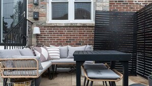 Un espace patio avec une table et des chaises noires, un canapé en osier avec des coussins et un mur de briques avec une fenêtre.