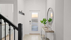 A hallway with a white door, a 'Welcome' sign, a mirror with beads, and a potted plant.