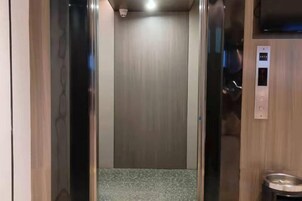 Interior de un ascensor con puerta metálica, panel de control y un pequeño cubo de basura.