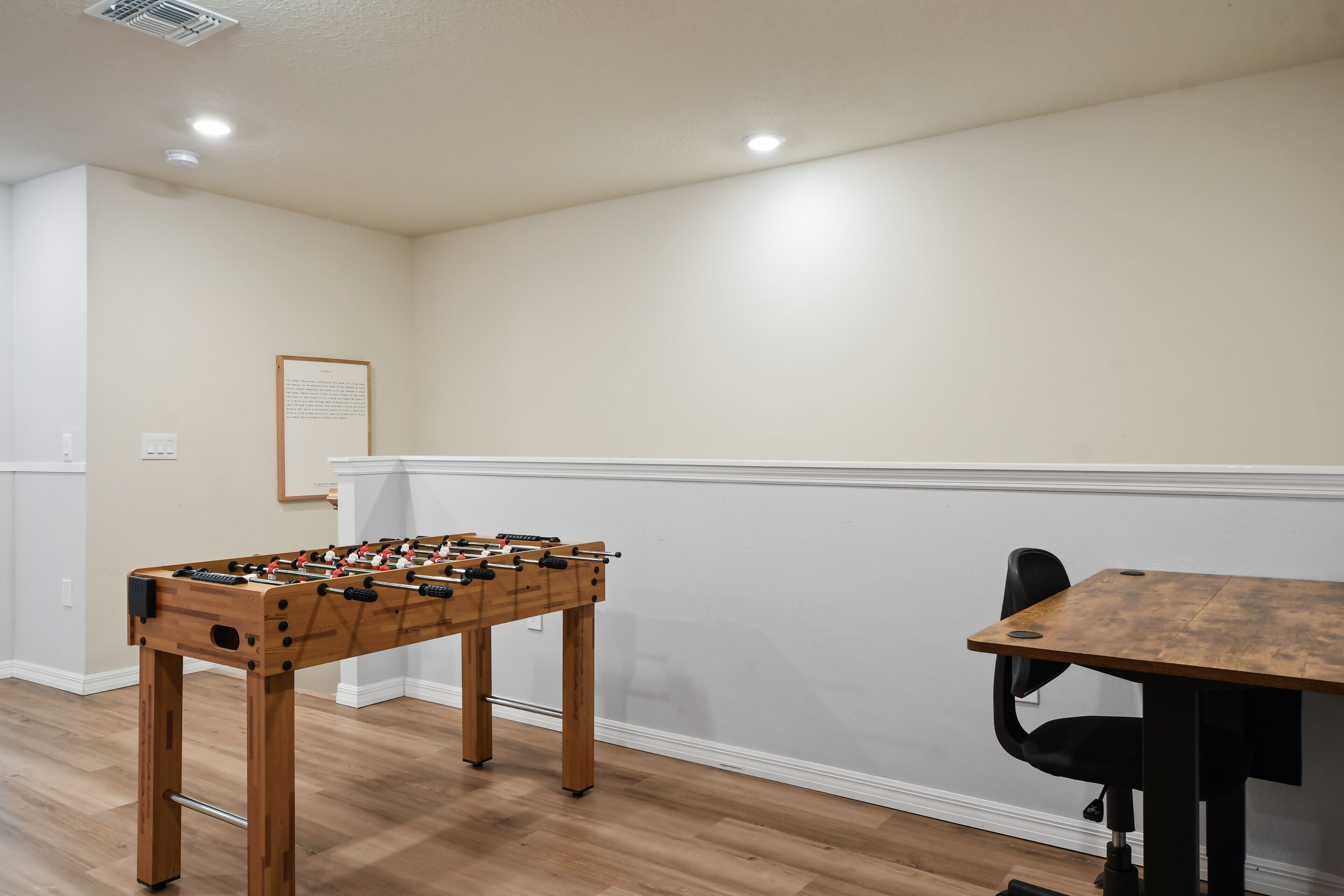 A room with a wooden foosball table, a white wall with a framed notice, and a dining table with chairs.