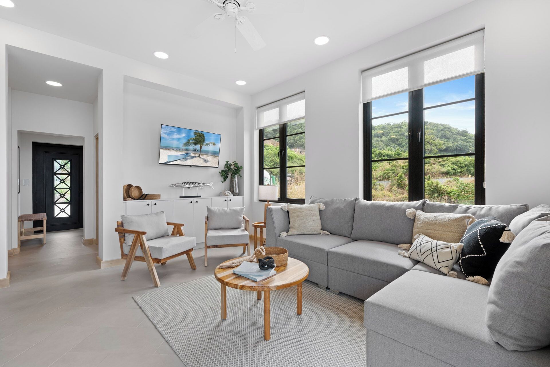 A spacious living room with a grey sofa, a wooden coffee table, and a TV mounted on the wall.