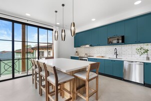 A modern kitchen with teal cabinets, a white countertop, and a dining area with wooden chairs.