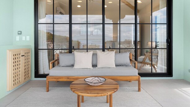 Et værelse med en stor skydedør, der fører til en balkon, en træsofa med puder, et rundt træbord med en flettet kurv og et træskab med et gittermønster.