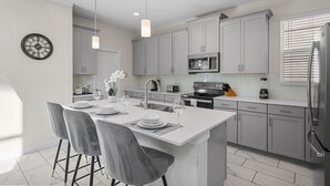 A modern kitchen with grey cabinets, a central island with seating, a clock on the wall, and a refrigerator.