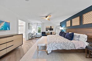 A bedroom with a bed, bedside tables, a desk, a chair, a ceiling fan, and artwork on the walls.