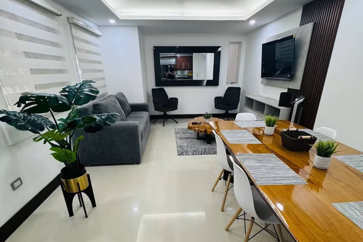 Ein modernes Wohnzimmer mit einer grauen Couch, einem Holzesstisch mit Stühlen und einem durch eine offene Tür einsehbarer Küchenbereich.