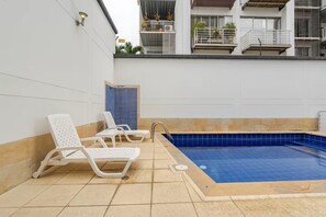 An outdoor swimming pool with blue tiles, surrounded by beige tiles and white walls. There are white lounge chairs and a blue shower area. - Edificio Fence Real (Cali)