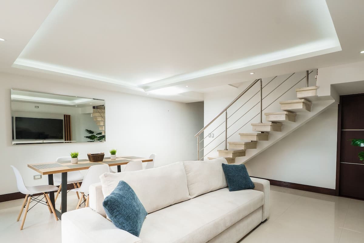 Ein modernes Wohnzimmer mit einer Couch, einem Esstisch mit Stühlen, einer Treppe und einem großen Spiegel an der Wand.