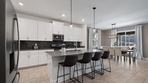 Une cuisine moderne avec des meubles blancs, un îlot central doté de tabourets, un coin repas avec une table et des chaises, et un éclairage suspendu.