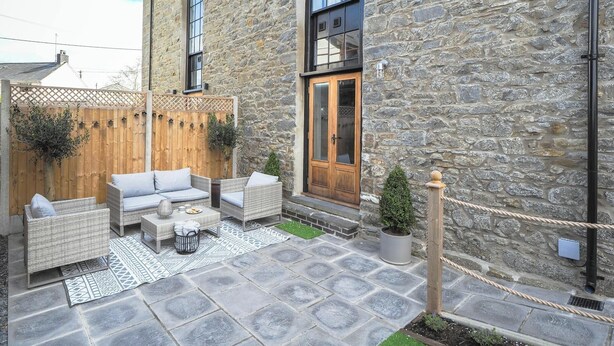 Un bâtiment en pierre avec une porte en bois, un petit jardin avec une table et des chaises, et une clôture en bois.