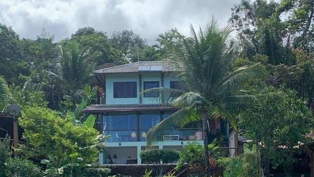 A two-story building with a balcony, surrounded by lush greenery and a body of water.