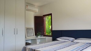 A bedroom with a bed, a wardrobe, a small table with a plant, and a window with a view of greenery.