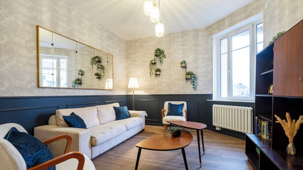 Una sala de estar con sofá, sillón, mesa de centro y estantería. Hay un espejo grande, plantas colgantes en la pared y un radiador.