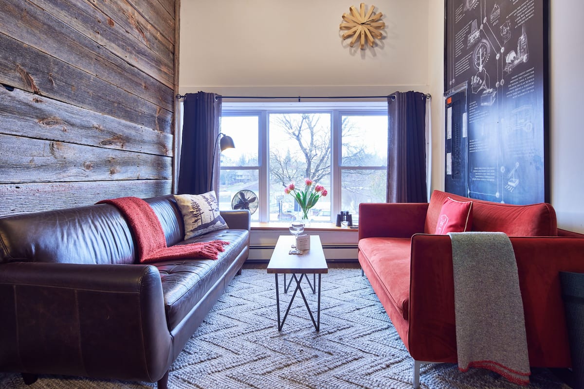 Una sala de estar con un sofá de cuero marrón, un sofá rojo, una mesita y una ventana con cortinas.