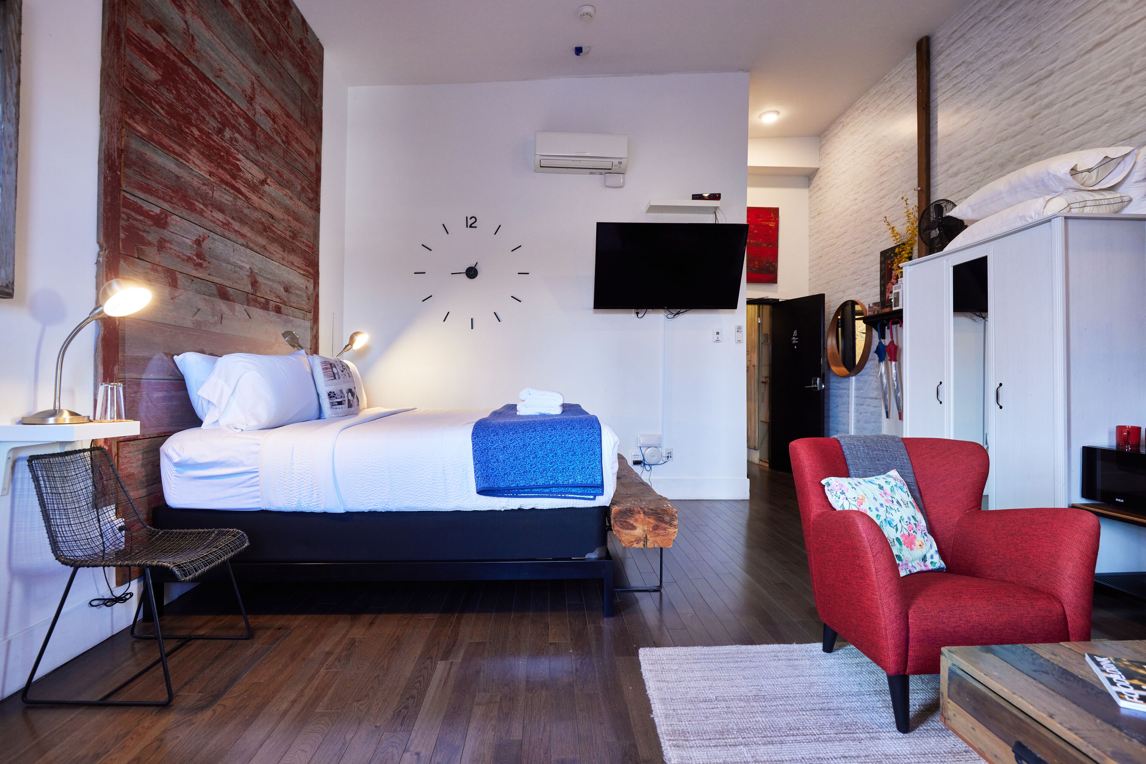A modern bedroom with a bed, a red armchair, a desk with a chair, a flat-screen TV, and a wall clock.
