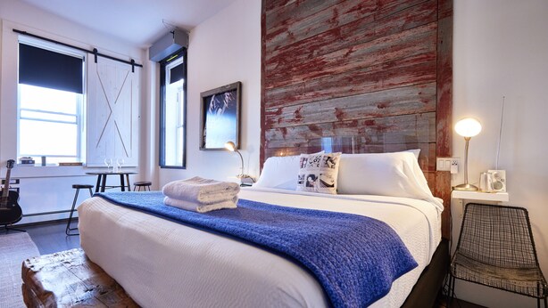 A neatly made bed with a blue blanket, a wooden headboard, and a wall-mounted picture. - Edisons Inn (Stratford)