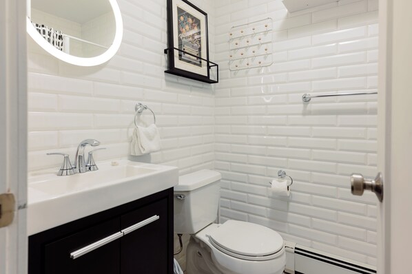 A modern bathroom with white subway tiles, a white sink with chrome fixtures, a toilet, and a framed picture above the sink.