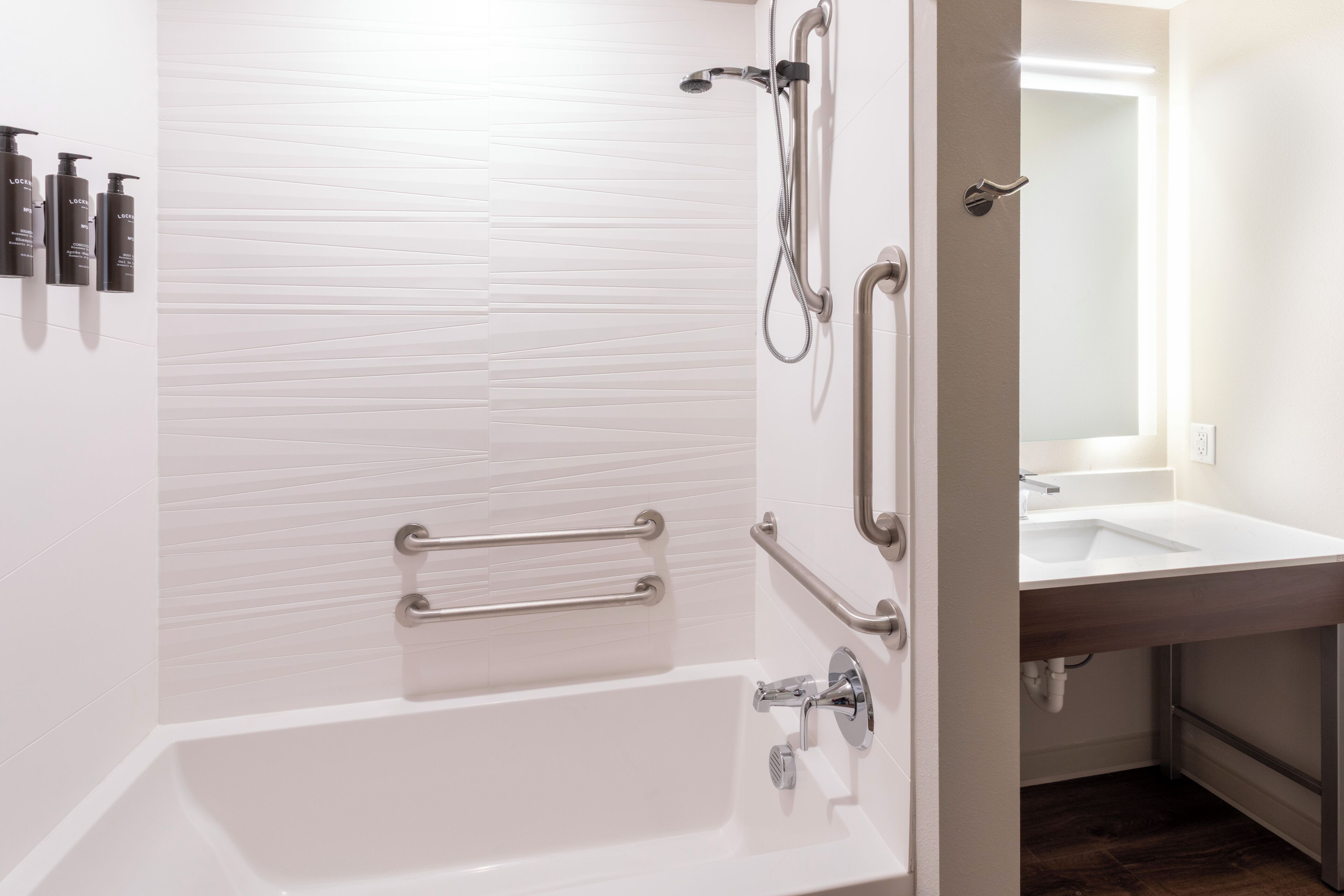 A modern bathroom with a white bathtub, a shower with grab bars, and a sink with a mirror above it.