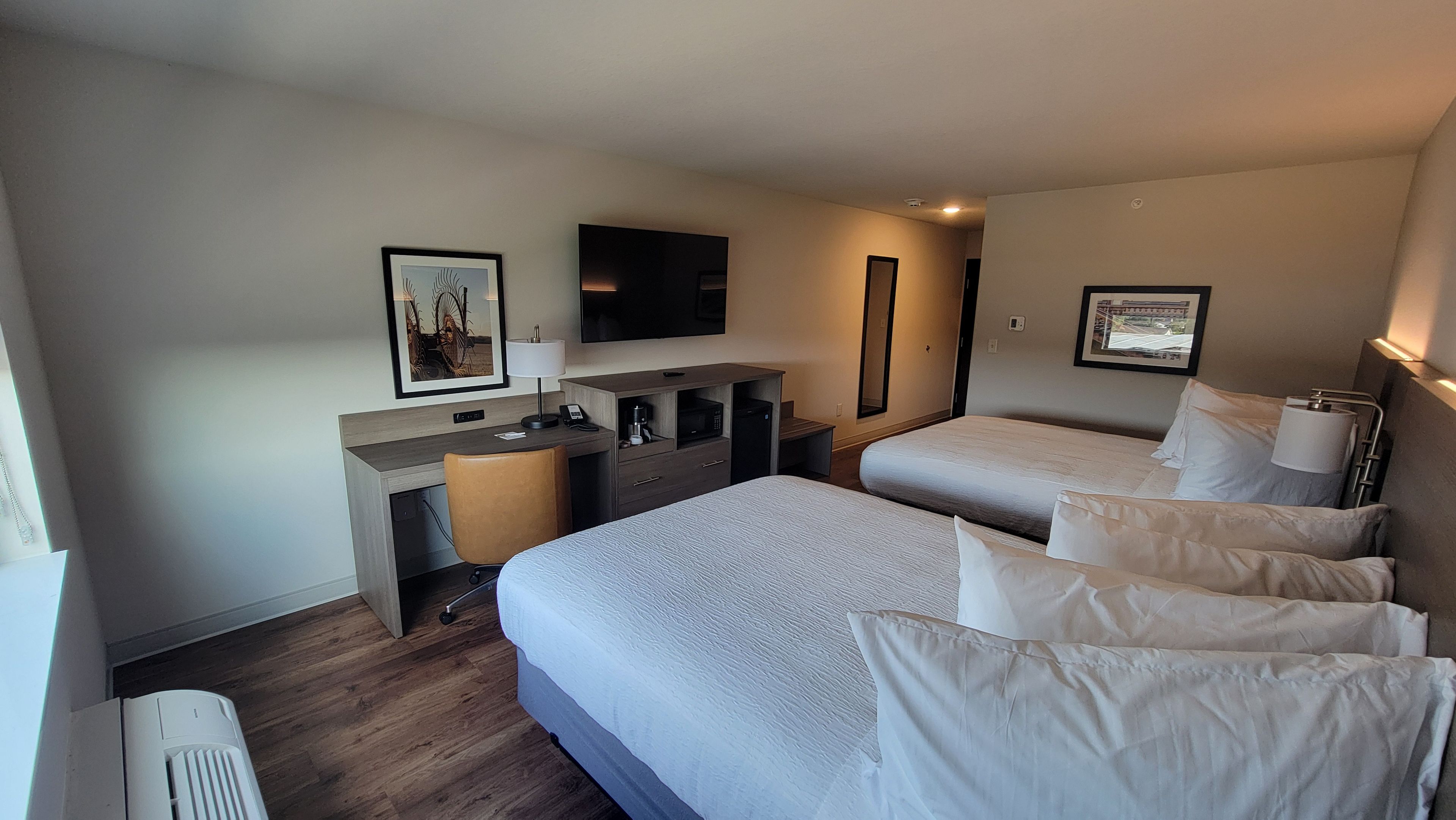 Quarto de hotel com duas camas, uma escrivaninha com cadeira, televisão de tela plana e um quadro na parede.