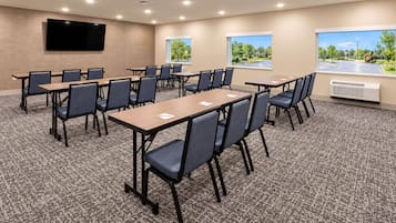 A conference room with multiple tables and chairs, a flat-screen TV mounted on the wall, and windows with a view of a parking lot and greenery.