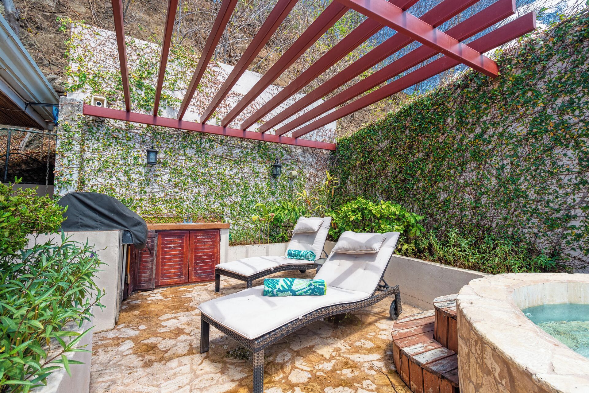 A sunlit outdoor patio with two lounge chairs, a small barbecue grill, and a stone-tiled floor.