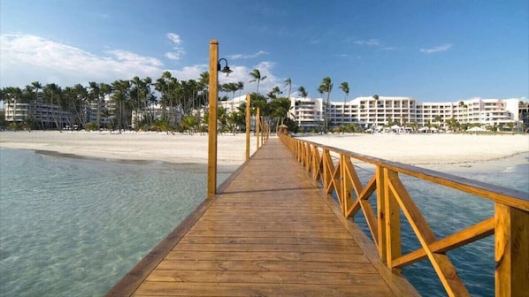 A wooden pier leading to a beach with clear water, palm trees, and hotel buildings in the background.