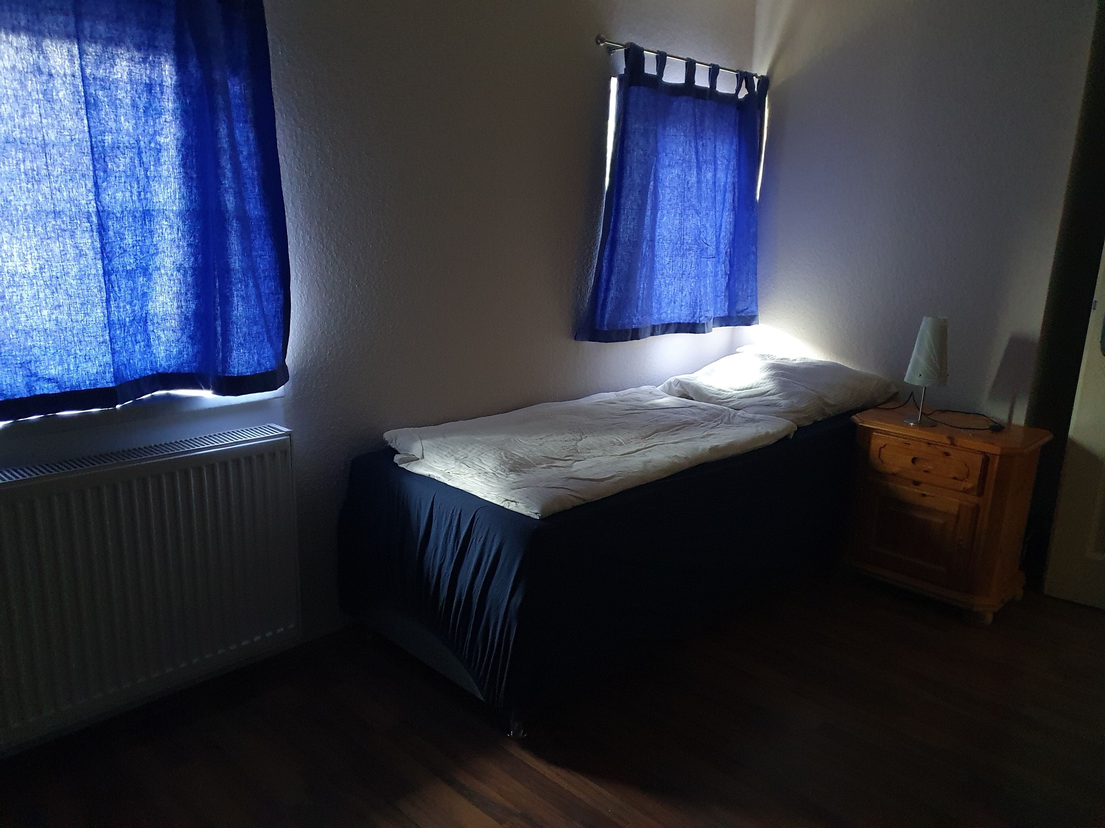 A small, dimly lit room with a single bed, a wooden nightstand, and a radiator.