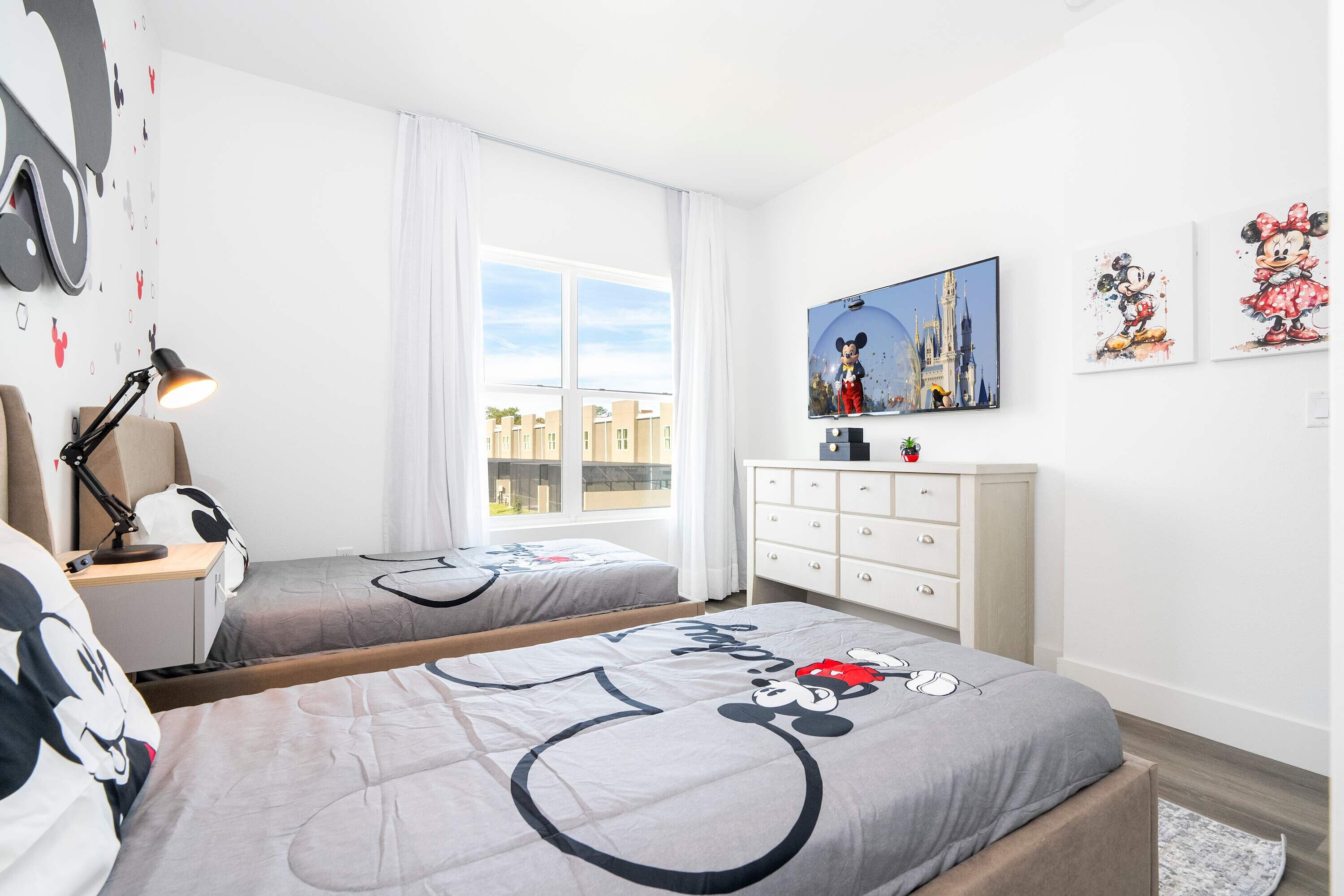 Habitación de hotel con dos camas, un escritorio, una lámpara, un aparador y ropa de cama con temática de Mickey Mouse.
