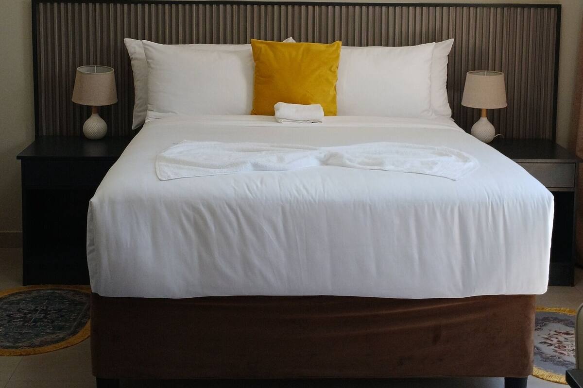 A neatly made bed with white and yellow pillows, a dark headboard, and a round ceiling light.