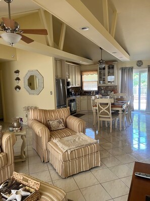 A spacious living area with a striped sofa, ottoman, and dining table with chairs. A kitchen with cabinets and a refrigerator is visible in the background. - Montauk 3BR/2BA w/ Multi-Level Decks Near Beach (Montauk)