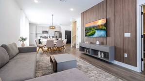 A modern living room with a gray sofa, a coffee table, and a TV mounted on a wooden wall. There is a dining area with a table and chairs in the background.