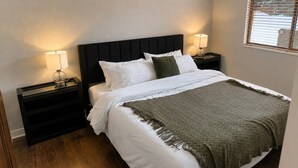 A neatly made bed with white linens and a green throw blanket, flanked by two bedside tables with lamps, in a room with wooden flooring and a plain wall. - The Powder Haven Chalet (Park City)