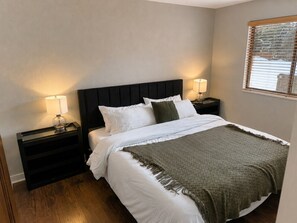A neatly made bed with white linens and a green throw blanket, flanked by two bedside tables with lamps, in a room with wooden flooring and a plain wall. - The Powder Haven Chalet (Park City)