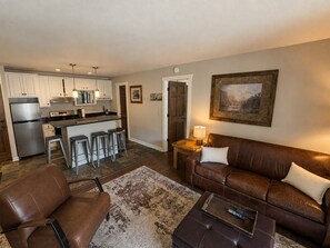 A living room with a brown leather sofa, a coffee table, and a rug. There is a kitchen area with white cabinets and a stainless steel refrigerator. A painting hangs on the wall. - The Powder Haven Chalet (Park City)