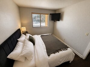 A bedroom with a bed, a nightstand, a lamp, a TV mounted on the wall, and a window with blinds. - The Powder Haven Chalet (Park City)