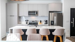 A modern kitchen with white cabinetry, a stainless steel refrigerator, a microwave, and a coffee machine. There are four bar stools with wooden legs and a white table with a black countertop. - Disney Getaway! Private Pool & Themed Bedrooms. Book Now! #DW36 (Orlando)