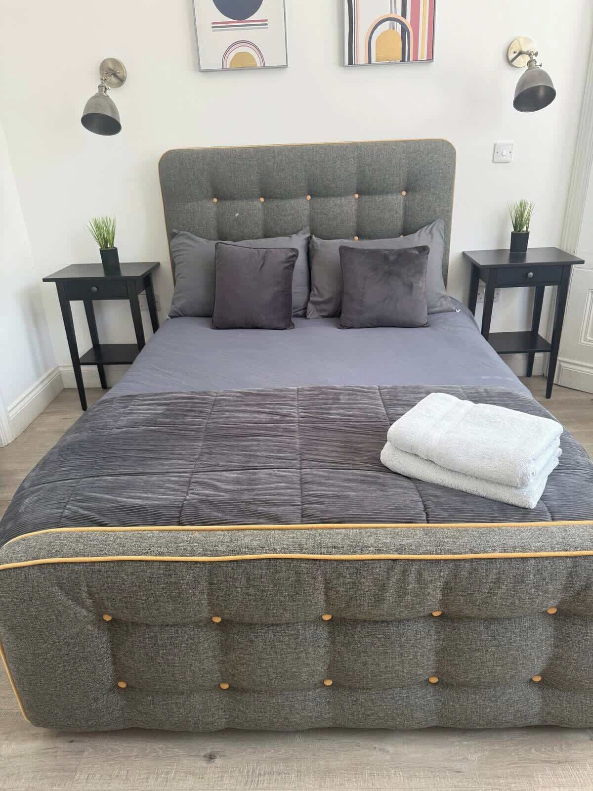 Una cama bien tendida con un tablero de cabecera y piecera tapizados en gris, flanqueada por dos mesitas de noche con lámparas.