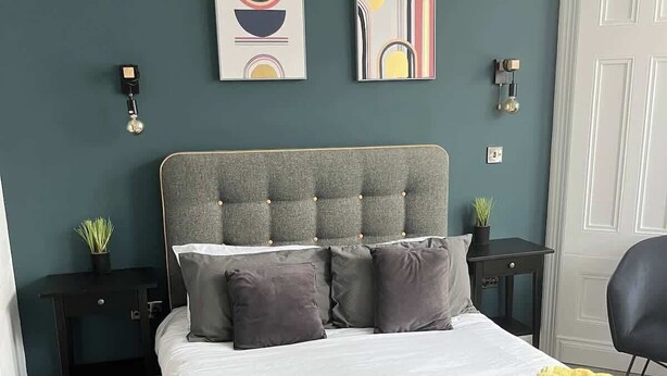 Une chambre à coucher avec une tête de lit capitonnée, un lit avec une literie grise, deux tables de chevet avec des lampes et des œuvres d'art abstraites accrochées au mur.